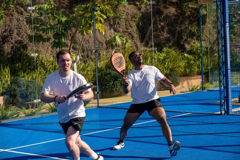 Two men playing tennis on a blue outdoor court with a hillside and fence in the background