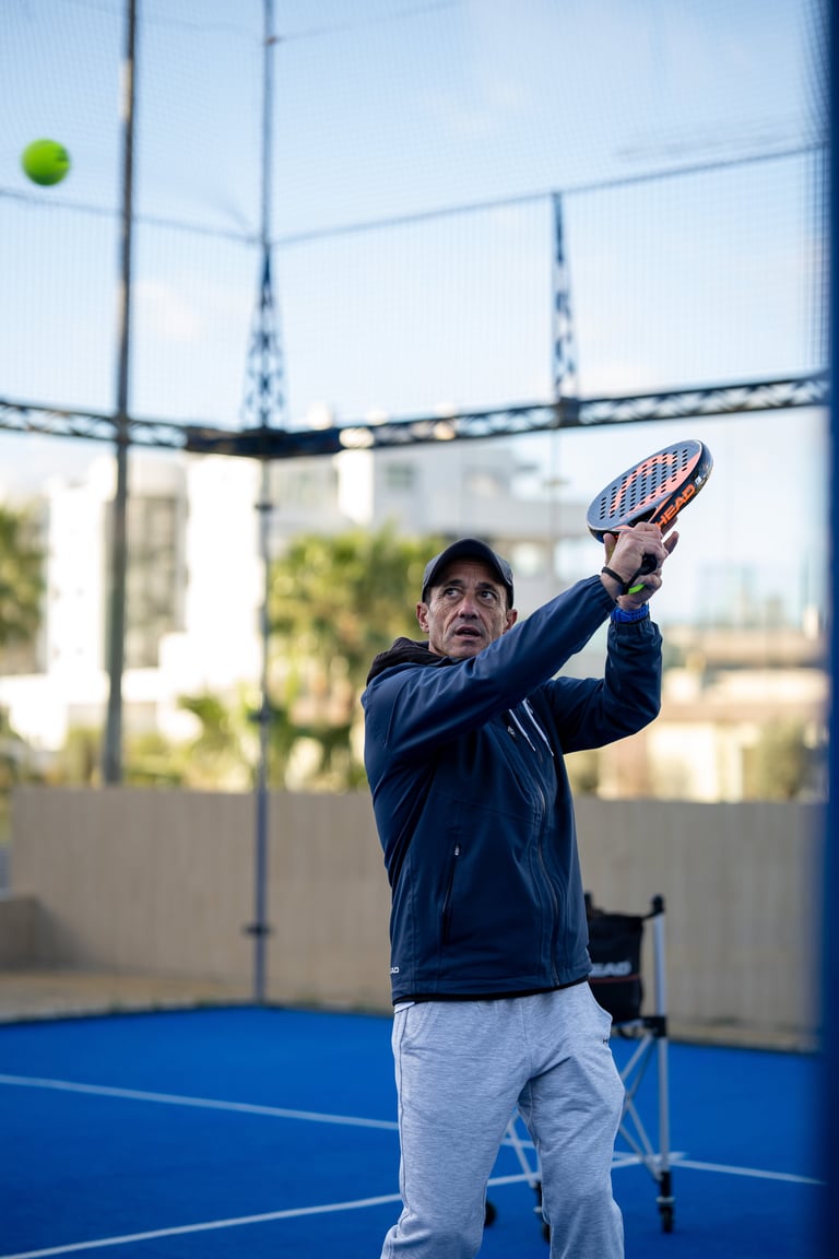 Man in blue jacket serving a tennis ball on an indoor blue court with a ball visible above